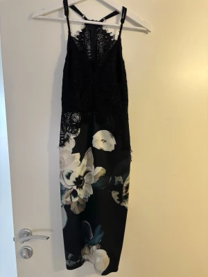 Svart spetsklänning med blommigt mönster - Snygg svart klänning med tunna axelband och spets upptill. Kjoldelen har ett stort blommigt mönster i vitt och beige på svart botten. Överdelen är i genombruten spets och klänningen har en figurnära passform. Perfekt för dig som vill sticka ut.