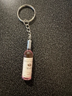 Nyckelring med vinflaska i miniatyr - Säljer en unik nyckelring med en miniatyrflaska vin som dekoration. Flaskan är rosa med en detaljerad etikett och sitter fast i en metallkedja och ring. Perfekt accessoar för dig som gillar vin och vill ha något personligt på nyckelknippan.