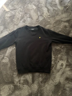 Svart sweatshirt med lyle & scott - Klassisk svart sweatshirt med rund hals och en liten gul broderad logga på bröstet. Tröjan har långa ärmar och ribbade muddar vid ärmslut och nederkant. Perfekt för en avslappnad och stilren look.