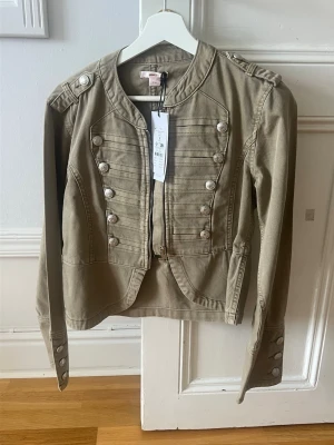 Grön jacka - Snygg olivgrön jacka med militärinspirerade detaljer, dekorativa silverknappar framtill och på axlarna. Jackan har lång ärm, markerade axlar och dragkedja framtill. Tillverkad i bomull för en bekväm känsla och cool look.