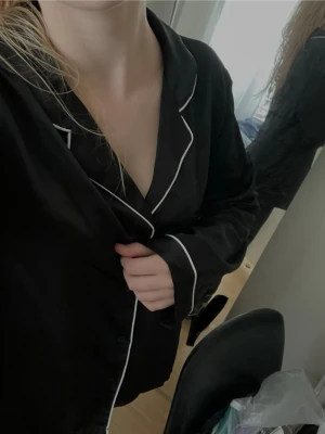 Svart pyjamas med vita kanter - Säljer en svart pyjamas med klassisk skjortmodell och vita kontrastsömmar. Setet består av långärmad överdel och långa byxor, båda i mjukt och glansigt material som känns lyxigt mot huden. Perfekt för mysiga kvällar hemma.