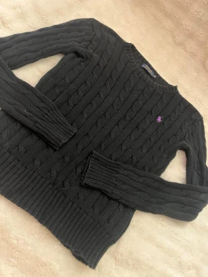 Svart kabelstickad tröja Polo Ralph Lauren - Kabelstickad svart tröja från Polo Ralph Lauren med lila broderad logga på bröstet. Tröjan har rund halsringning, långa ärmar och ribbade muddar vid ärmslut och nederkant. Perfekt för dig som gillar klassisk och stilren design.