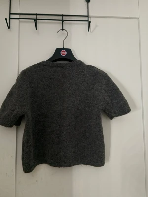 Mörkgrå stickad t-shirt - Mysig mörkgrå stickad t-shirt med korta ärmar och rund hals. Tröjan har en enkel och stilren design, perfekt för lager på lager och passar till många olika outfits. Materialet ser ut att vara mjukt och lite luddigt, vilket ger en avslappnad vibe.