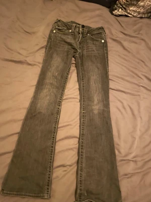 Grå bootcut jeans från True Religion - Säljer ett par grå bootcut jeans från True Religion gmed snygga detaljer på bakfickorna och silverfärgade knappar. Jeansen har låg midja och klassisk femficksdesign. Perfekta för dig som gillar en avslappnad men trendig stil.