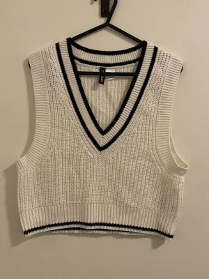 Vit stickad väst med svarta detaljer - Vit stickad väst med djup V-ringning och svarta ränder längs halsringning och nederkant. Croppad modell i grovstickat material, perfekt att styla över en skjorta eller t-shirt. Klassisk och trendig look med sportig känsla.