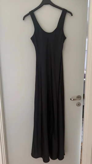 Svart långklänning i satin - En elegant svart långklänning med smala axelband och rundad urringning. Klänningen har en slät, glansig yta som ger en lyxig känsla och ett stilrent fall. Perfekt för dig som vill ha en enkel men snygg look.