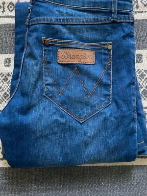Blå bootcut jeans från Wrangler - Ett par klassiska blå bootcut jeans från Wrangler. Med de ikonisk W-detalj på bakfickan. Midjan är lowwaisted/midwaisted, men sitter ändå lågt vid höfterna. Midjemåttet rakt över är 37,5 cm och inre benlängden är 78,5 cm.