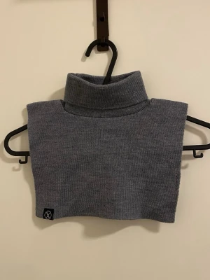Grå stickad krage i mjukt material - Säljer en grå stickad krage med polokrage, perfekt att bära under jackan för extra värme. Kragen är i mjukt och stretchigt material och har en enkel, stilren design. Liten svart logga på kanten framtill. Passar dig som vill ha något både praktiskt och snyggt.
