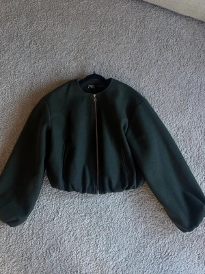 Mörkgrön cropped jacka från Zara - Snygg mörkgrön cropped jacka från Zara med rund halsringning och dragkedja framtill. Jackan har ballongärmar och resår i nederkanten för en modern siluett. Tillverkad i ett tjockare, filtliknande material som passar perfekt till höst och vinter.