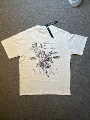 Vit AMIRI t-shirt med ängeltryck - Säljer en vit t-shirt från AMIRI med stort grafiskt tryck av en ängel och moln på ryggen. T-shirten har korta ärmar och en relaxed passform. Perfekt för dig som gillar streetwear och unika prints.