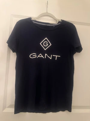 Mörkblå GANT t-shirt med logga - Snygg mörkblå t-shirt från GANT med vit logga och tryck på bröstet. Klassisk rund halsringning och korta ärmar. Perfekt till jeans eller shorts för en enkel och stilren look.
