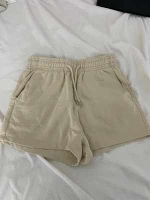 Beige mjuka shorts med snörning - Beiga shorts i mjukt material med elastisk midja och snörning. De har två sidofickor och avslappnad passform, perfekta för varma dagar eller chill hemma. Klassisk och enkel design som är lätt att matcha.