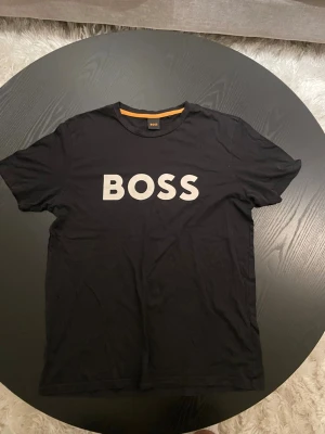 Boss t-shirt - Säljer en svart t-shirt från BOSS med stort vitt BOSS-tryck på bröstet. Klassisk rund halsringning och korta ärmar. Tillverkad i mjuk bomull för skön känsla och enkel stil.
