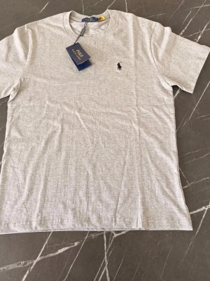 Ljusgrå t-shirt från Polo Ralph Lauren - Snygg ljusgrå t-shirt från Polo Ralph Lauren med klassisk passform och liten broderad logga på bröstet. Tillverkad i mjuk bomull som känns skön mot huden. Perfekt basplagg till garderoben.