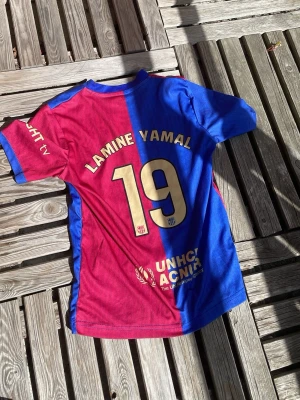 Blåröd Lamine Yamal matchtröja - Säljer en blåröd fotbollströja med Lamine Yamal och nummer 19 på ryggen. Tröjan har klassisk halv-halv design i rött och blått, rund hals och tryck i guld. Materialet är lätt och andas, perfekt för träning eller match. UNHCR-logga på nedre delen av ryggen.