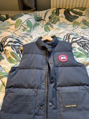 Mörkblå dunväst från Canada Goose XS - mörkblå dunväst från Canada Goose med hög krage och två stora fickor framtill. Västen har det klassiska Canada Goose-märket på bröstet och stängs med både dragkedja och knappar. Xs