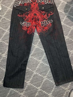 Mörkgrå jeans med röd tryck - Unika mörkgrå jeans med rak passform och stort rött tryck på baksidan med text och grafiskt mönster. Jeansen har två bakfickor med knapp och detaljerade sömmar. Materialet är klassisk denim som ger en cool streetwear-vibe.