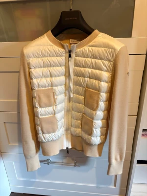 Beige och vit cardigan från Moncler - Snygg cardiganjacka från Moncler i beige och vitt. Framsidan är quiltad och vadderad med två stickade fickor, medan ärmar och rygg är i mjuk stickad ull. Dragkedja framtill och Moncler-logga på ärmen. Perfekt för lager-på-lager och en trendig look. Självklart äkta och sparsamt använd!