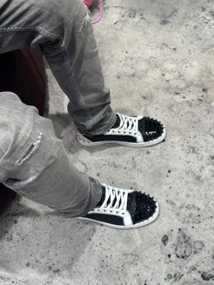 svarta sneakers - Unika sneakers i svart och vitt med glittrande detaljer och svarta nitar på tån. Skorna har vita snören, vit sula och den klassiska röda undersidan. Perfekt för dig som vill sticka ut med din stil.