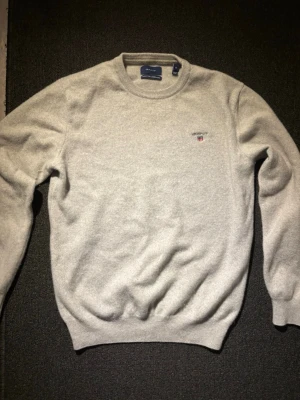 Gant sweatshirt  - Supermjuk ljusgrå stickad tröja från Gant i lammull. Klassisk rund halsringning och ribbade muddar. Liten broderad Gant-logga med sköld på bröstet. Perfekt för lager på lager och riktigt skön att ha på sig.