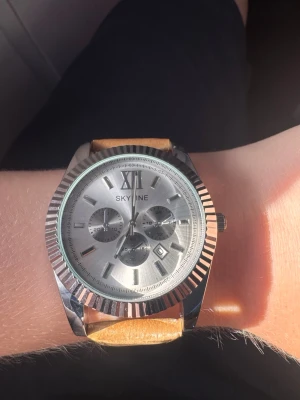 Silverfärgad Skyline klocka med läderband - Stilren klocka från Skyline med rund silverfärgad urtavla och markerade index. Klockan har flera små urtavlor och datumvisning. Armbandet är i brunt läder som ger en snygg kontrast till det blanka metallboetten. Ingen låda