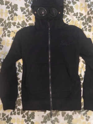 Svart hoodie med goggles från C.P. Company - Svart hoodie från C.P. Company med hel dragkedja och ikoniska goggles i huvan. Hoodien har långa ärmar, två stora fickor framtill och är tillverkad i mjukt material. Perfekt för dig som vill sticka ut med en unik streetwear-look. Aldrig använt eftersom den var för stor på mig. Storleksguide kan skickas om önskad.