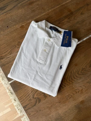 Vit pikétröja från Polo Ralph Lauren - Klassisk vit pikétröja från Polo Ralph Lauren med korta ärmar och krage. Tröjan har två knappar framtill och den ikoniska broderade loggan på bröstet. Perfekt till jeans eller shorts för en clean och stilren look.