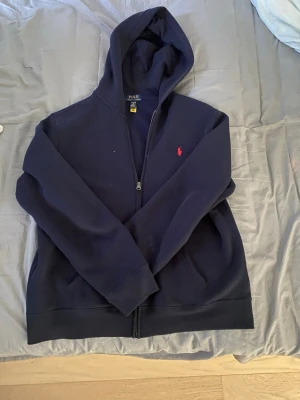 Mörkblå hoodie från Polo Ralph Lauren - Mörkblå hoodie med dragkedja från Polo Ralph Lauren. Klassisk design med huva, två fickor framtill och den ikoniska röda broderade loggan på bröstet. Tillverkad i mjukt bomullsmaterial som är skönt att bära.