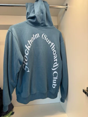 Stockholm Surboard Club hoodie - Säljer en riktigt snygg blå hoodie från Stockholm Surfboard Club i stl xs men passar också liten s. Köpt i butik för 3000kr och varsamt använd. Tröjan är i topp skick förutom ett litet hål vid fickan som inte syns vid användning.