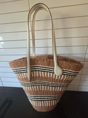 Randig shoppingväska i strå - Snygg shoppingväska i naturfärgat stråmaterial med vita och svarta ränder. Väskan har långa, vita handtag i konstläder och en rymlig, konformad design. Perfekt för att bära allt du behöver till skolan, stranden eller stan.