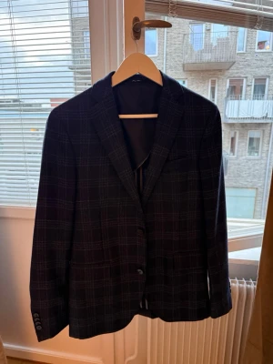Mörkblå rutig kavaj Mango - Snygg mörkblå kavaj med diskret rutmönster. Klassisk modell med två knappar framtill, smala slag och fickor. Perfekt för dig som vill ha en stilren och modern look. Materialet ser ut att vara en blandning med ullkänsla.