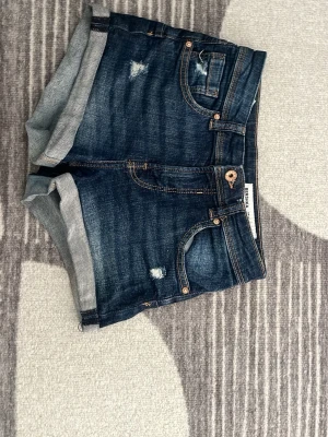 Mörkblå denimshorts med upprullad kant - Säljer ett par mörkblå jeansshorts med upprullad kant och slitna detaljer. Klassisk femficksmodell med knapp och dragkedja framtill. Perfekta för sommaren och ger en avslappnad vibe.