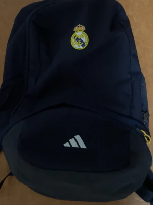Mörkblå Real Madrid ryggsäck Adidas - Snygg mörkblå ryggsäck med Real Madrid-logga och Adidas-märke på framsidan. Ryggsäcken har flera fack, meshficka på sidan och dragkedja med gul detalj. Perfekt för skola eller träning och tillverkad i slitstarkt tygmaterial. Helt ny använd 1 gång priset kan diskuteras , säljer pga har jätte många väskor och vill bli av med just den och andra väskor kommer 😁😉