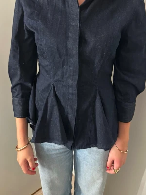 Mörkblå figursydd skjorta - Snygg mörkblå skjorta i denim material med figursydd passform och markerad midja. Skjortan har långa ärmar, dolda knappar framtill och veckade detaljer nedtill som ger en unik siluett. Perfekt för dig som vill ha en stilren men ändå cool look.