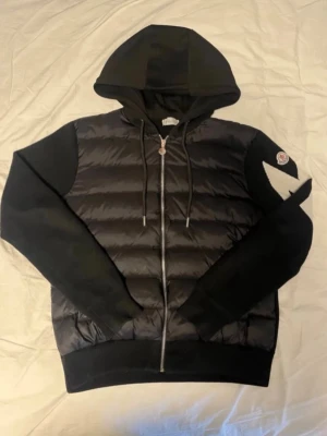 Svart Cardigan - Snygg svart pufferjacka med huva och dragkedja framtill. Jackan har quiltad kropp i glansigt material och stickade ärmar samt muddar. På vänster ärm finns en vit detalj och en rund logotyp. Perfekt för kalla dagar och riktigt trendig look.