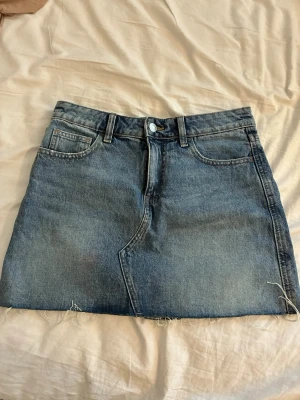 Helt ny kort jeanskjol - Snygg kort jeanskjol i blå denim med råa kanter och broderade detaljer på bakfickorna. Kjolen har klassisk femficksdesign, bälteshällor och knappstängning framtill. Storlek 34 från Gina tricot. Helt ny. 