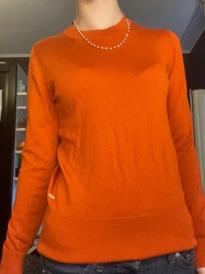 Orange stickad tröja med knappar - Snygg orange stickad tröja med rund hals och långa ärmar. Tröjan har dekorativa guldfärgade knappar vid ärmsluten och ribbade muddar. Perfekt för dig som vill ha en färgklick i garderoben.