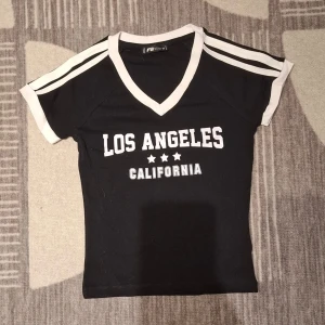 Svart v-ringad topp med text - Snygg svart topp med vit v-ringning och vita detaljer på ärmarna. Tryck med texten 'Los Angeles California' och tre stjärnor framtill. Kortärmad och figurnära passform, perfekt till jeans eller kjol.