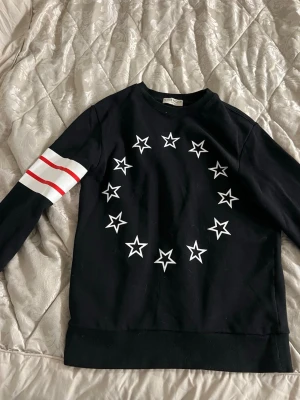 Svart sweatshirt från Givenchy - Svart sweatshirt från Givenchy med vita stjärnor i cirkel på bröstet och två röda ränder på ena ärmen. Tröjan har rund halsringning och ribbade muddar. Perfekt för dig som gillar streetwear och vill sticka ut med en exklusiv look. size S