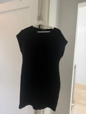 Svart ärmlös kortklänning - En enkel och stilren svart kortklänning med rak passform och ärmlös design. Perfekt basplagg som kan stylas på många olika sätt. Klänningen har rund halsringning och är tillverkad i ett mjukt material.