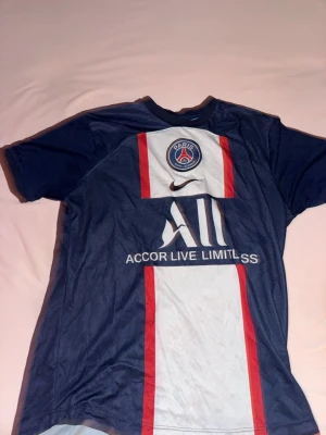PSG matchtröja Hakimi #2 blå - Säljer en blå PSG matchtröja från Nike med Hakimi och nummer 2 på ryggen. Tröjan har vita och röda detaljer, klubbmärke och sponsortryck på bröstet samt korta ärmar. Perfekt för dig som älskar fotboll och vill stötta Paris Saint-Germain.