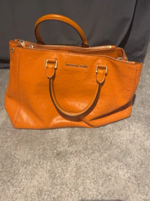 Orange handväska från Michael Kors - Snygg orange handväska från Michael Kors i präglat läder med struktur som påminner om struts. Väskan har två handtag, guldfärgade metalldetaljer och dragkedja upptill. Perfekt för dig som vill sticka ut med en färgglad accessoar.