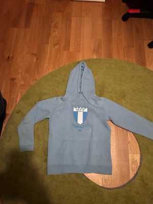 Ljusblå Malmö FF hoodie med huva - Säljer en ljusblå hoodie med Malmö FF-logga på bröstet. Hoodien har klassisk huva med dragsko och en stor magficka framtill. Perfekt för dig som vill visa ditt stöd för laget på ett avslappnat sätt.