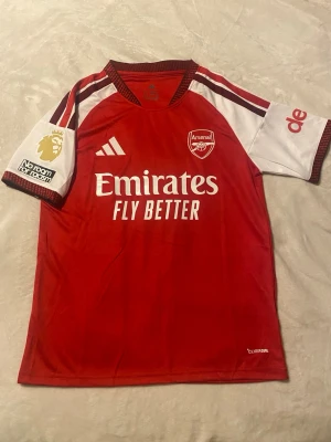 Arsenal matchtröja Saka 7 röd - Säljer en officiell Arsenal matchtröja från Adidas i rött och vitt med Saka 7 på ryggen. Tröjan har klubbmärke, sponsortryck och Premier League-märke på ärmen. Tillverkad i lätt och ventilerande funktionsmaterial, perfekt för fotbollsträning eller match. Helt ny! Pris kan sänkas vid snabb affär!