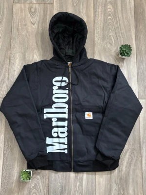 Svart Carhartt x Marlboro jacka - Snygg svart jacka med dragkedja från Carhartt, prydd med stor vit Marlboro-logga längs framsidan. Utrustad med huva och ribbade muddar. Praktisk ficka framtill med Carhartt-märke. Perfekt för dig som gillar streetwear och vill sticka ut.