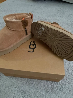 Beige UGG boots med dragkedja - Säljer ett par beige UGG boots i mocka med dragkedja på sidan och mjukt foder inuti. Skorna har en rejäl sula och klassisk UGG-logga på sidan. Perfekta för kalla dagar och riktigt sköna att ha på sig.