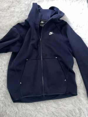 Mörkblå zip hoodie från Nike - Snygg mörkblå hoodie från Nike med dragkedja framtill och vit logga på bröstet. Tröjan har huva och stora fickor framtill. Tillverkad i mjukt bomullsmaterial som känns skönt mot huden. Perfekt för en avslappnad och sportig stil.