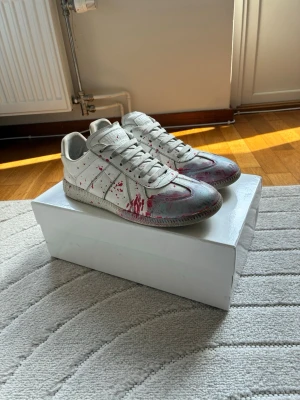 Maison margiela gats  - Säljer nu mina Maison margiela gats med röd och blå färgstänk, skorna är ganska unika då just denna kombination av färgstänk inte är så vanligt. Skorna är använda som man kan se men det har fortfarande mycket liv kvar, men där av det nedsatta priset, har du några frågor tveka inte att fråga!😁 