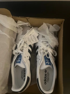 Adidas samba Originals vita sneakers - Snygga vita Adidas Originals sneakers med svarta ränder och klassisk blå logga på plösen. Skorna har en låg profil och gummisula i brunt. Tillverkade i läder med textildetaljer och vita skosnören. Perfekta för en clean och sportig stil.