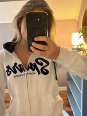 Vit Tottenham hoodie med dragkedja - Säljer en vit hoodie från Tottenham hotspur med dragkedja och mörkblå detaljer. Hoodien har huva med mörkblå snören och innerfoder samt två fickor framtill.